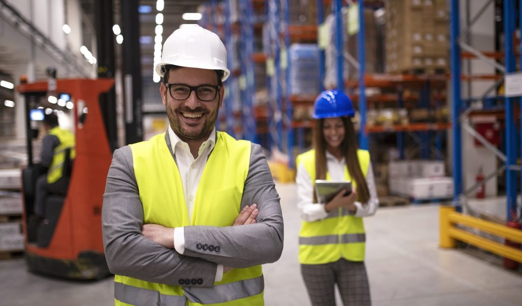 Portrait of successful warehouse worker or supervisor with crossed arms standing in large storage distribution area.